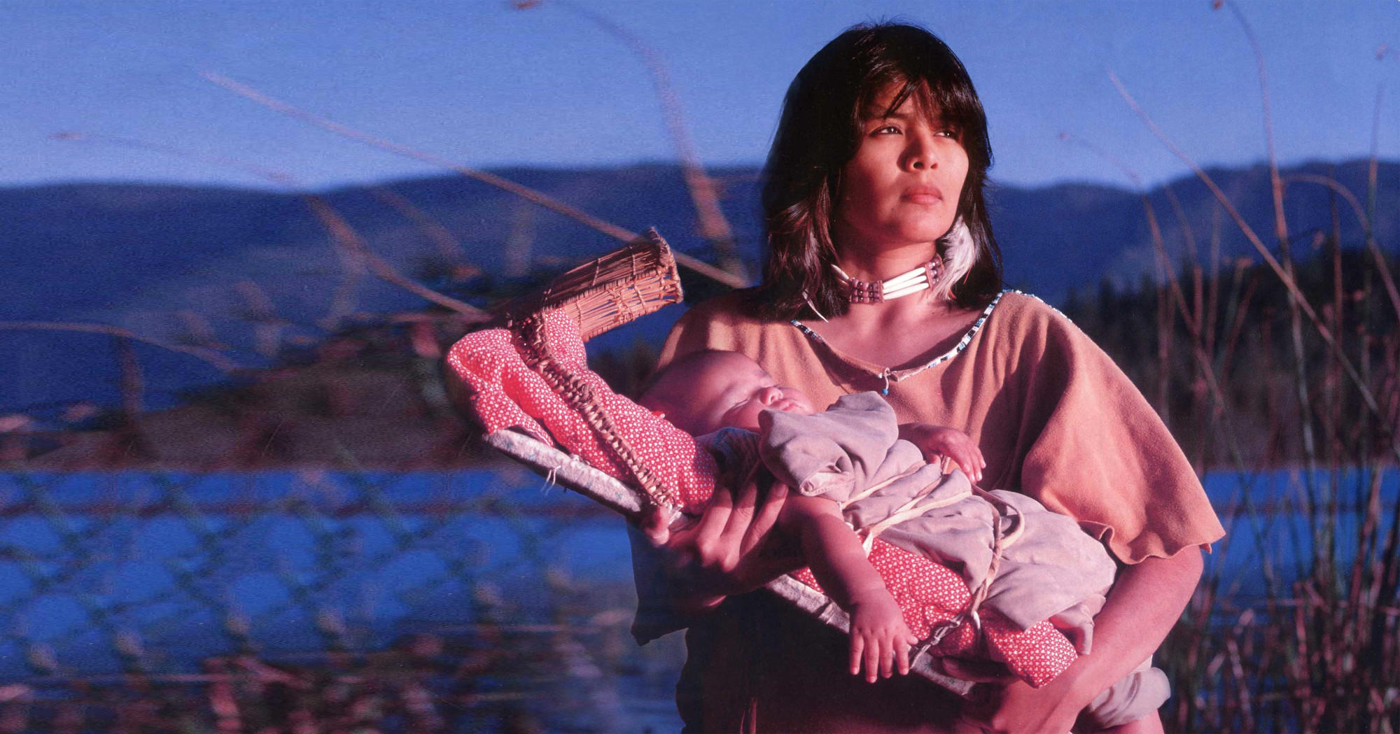 Native woman in traditional garb holding a baby and looking off into the distance